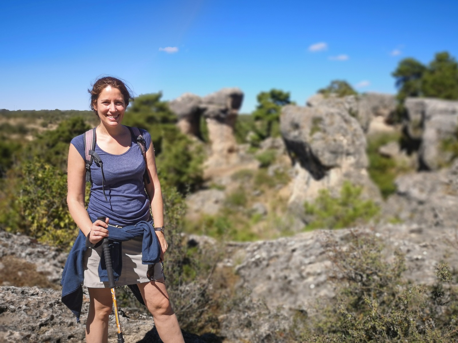 Lara está en la parte izquierda de la foto muy sonriente con un bastón de trekking y mochila. Al fondo un paisaje de montaña con rocas y vegetación.
