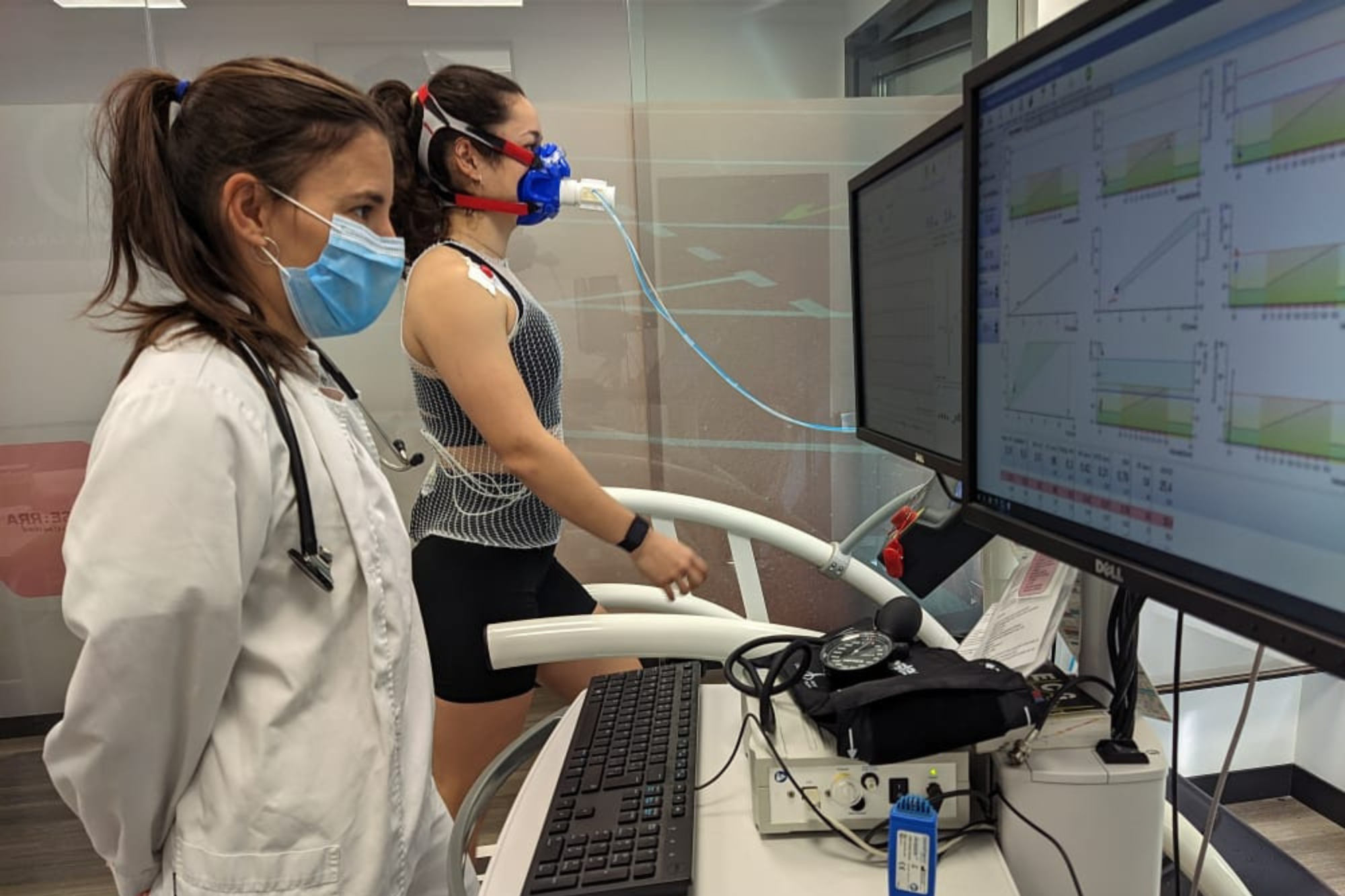 Anna aparece en primer plano a la izquierda de la foto en un laboratorio con una bata blanca de médico y visualizando unos monitores con datos. Detrás de ella, al fondo, se ve a una deportista en un tapiz rodante realizando una prueba de esfuerzo, con una máscara para medir gases.