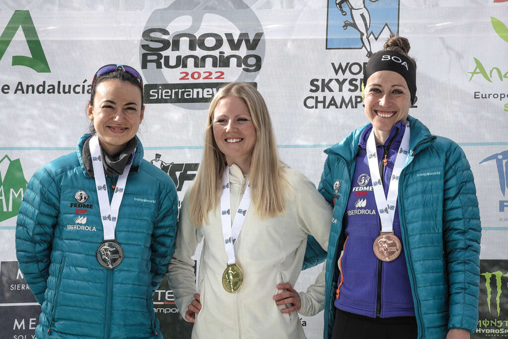 Tres mujeres sonrientes. Cada una con una medalla, detrás de una pancarta de Snow Running 2022