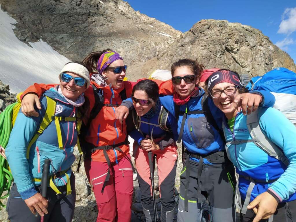 Imagen de un grupo de mujeres abrazadas, sonrientes, ventidas con ropa de montaña y el fondeo montañas, una de ellas con nieve