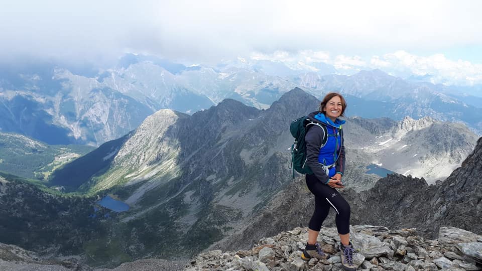 Elvira aparece de pie en primer plano, haciendo montañismo en Val de Aosta, al fondo las montañas y un mar de nubes espectacular. Lleva mallas negras, chaqueta azul y una mochila verde a la espalda. De su amplia sonrisa se desprende que está disfrutando de la experiencia.