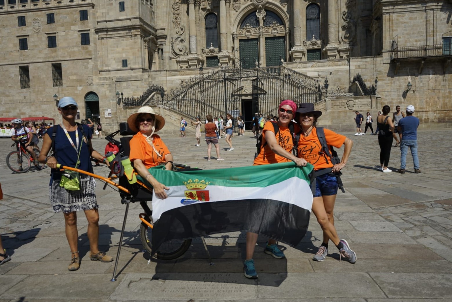 En la imagen se puede ver a Yolanda Álvarez, en la plaza del Obradoiro, frente a la Catedral de Santiago de Compostela. Está montada en una silla Joëlette y posa, exhibiendo la bandera de Extremadura (su tierra), junto a una parte del equipo de pilotos que hicieron posible su Camino de Santiago.