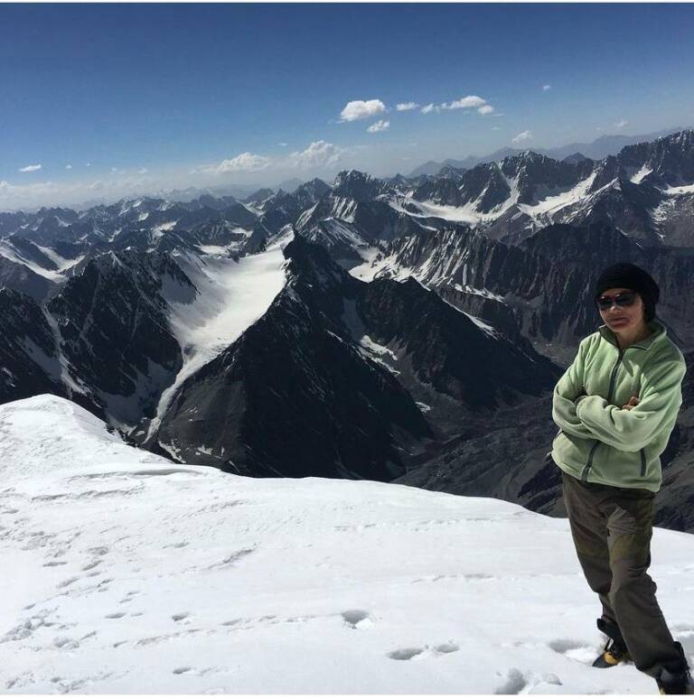Aqila Hashimi aparece en primer plano con vestimenta de montaña verde, gorro negro, gafas de sol y los brazos cruzados, sobre un llano de nieve. Tras de ella un espléndido paisaje de picos de roca y nieve en sus cumbres y corredores.