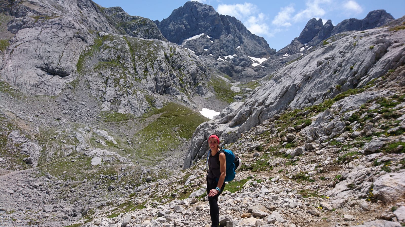 Noelia aparece en primer plano durante una jornada de senderismo en el Parque Nacional de los Picos de Europa. Va vestida con pantalón y camiseta de color negro, una cinta roja en su pelo y mochila en la espalda. Tras ella un impresionante paisaje de Picos, con roca, nieve y vegetación verde.