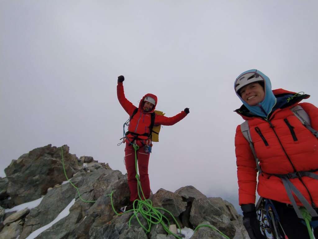 Dos mujeres alpinistas con ropa de abrigo en la cima de una montaña, con algo de nieve, en actitud de celebración