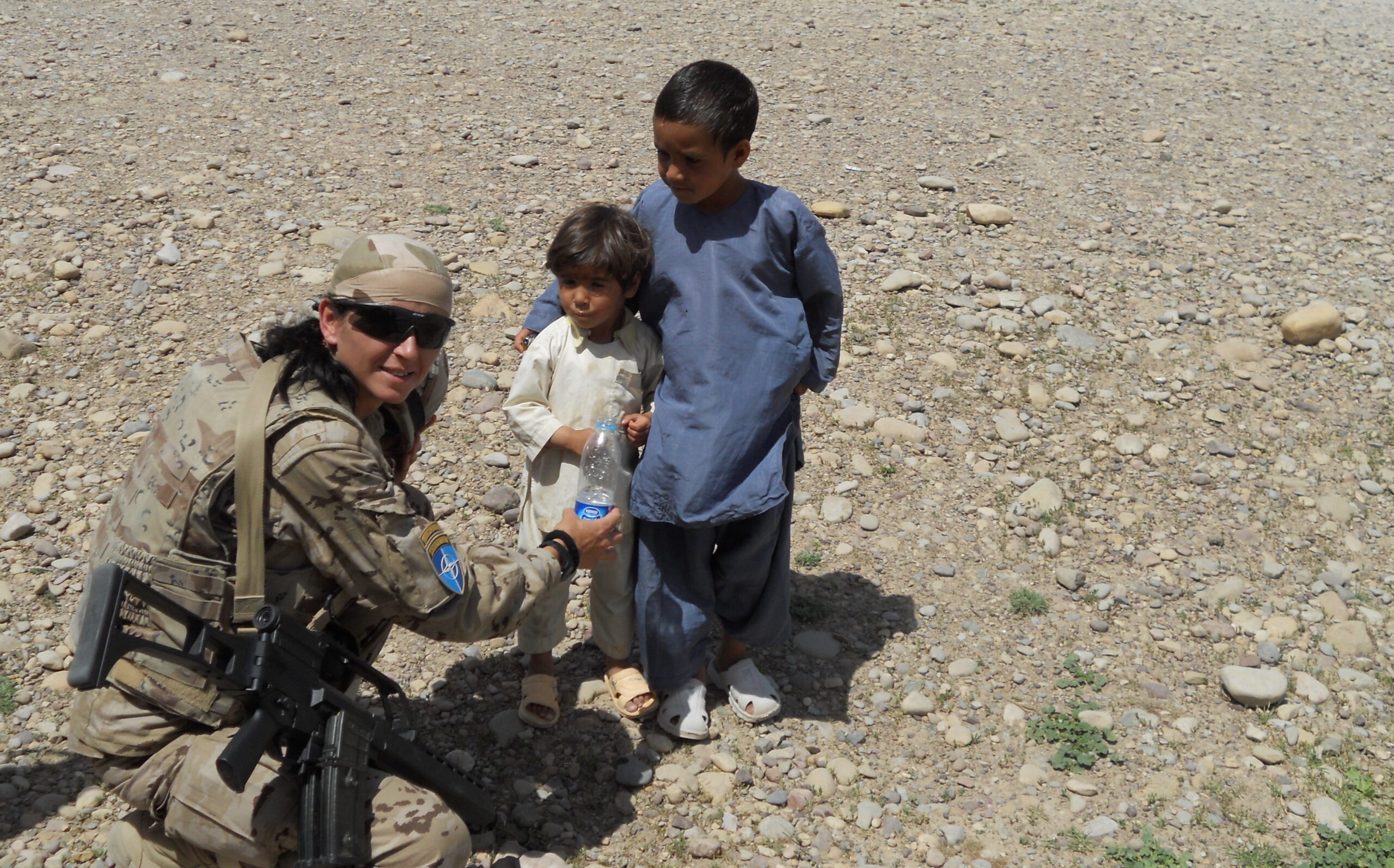La Cabo 1ª Adelina Torres aparece a la izquierda de la foto con uniforme de campaña, agachada ofreciendo una botella de agua a dos niños mientras patrulla el aeropuerto de Qala-e-Naw en Afganistán.