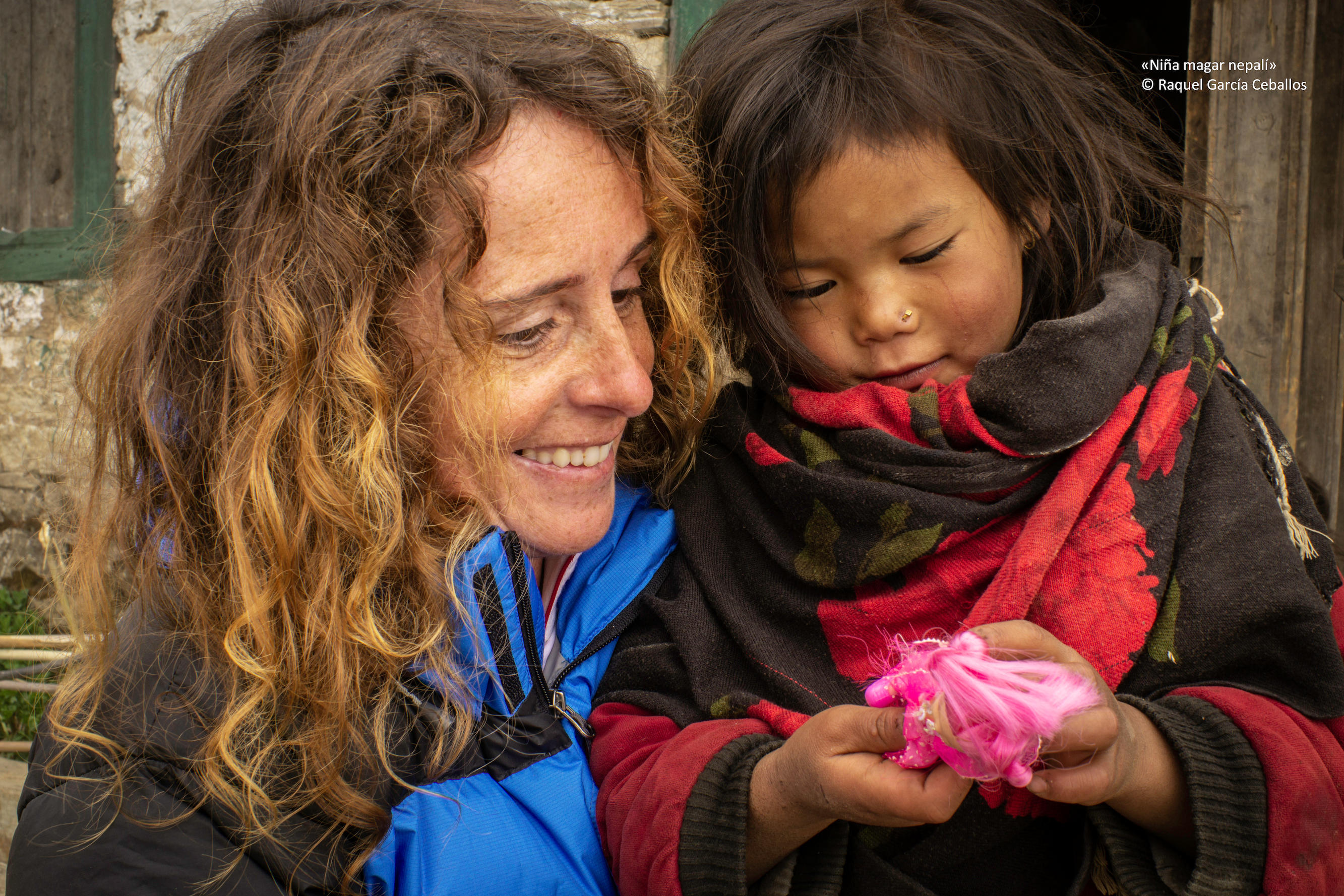Raquel aparece en la parte izquierda de la foto sosteniendo a una niña nepalí que aparece a la derecha y sostiene una muñeca con las manos. Es un primer plano y ambas sonríen observando la muñeca.