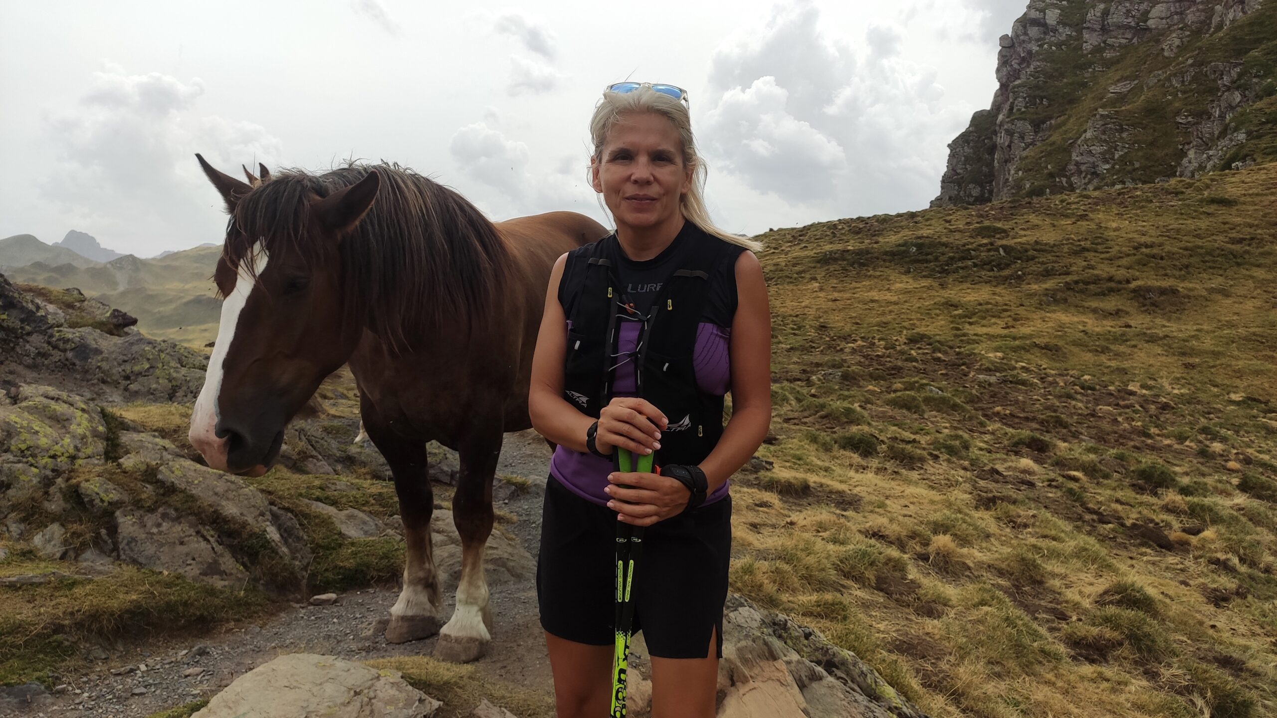 Peli aparece en medio de la foto en un paisaje montañoso con ropa de montaña y bastones de marcha nórdica. A su izquierda y justo detrás tiene un caballo que parece estar posando junto a ella.